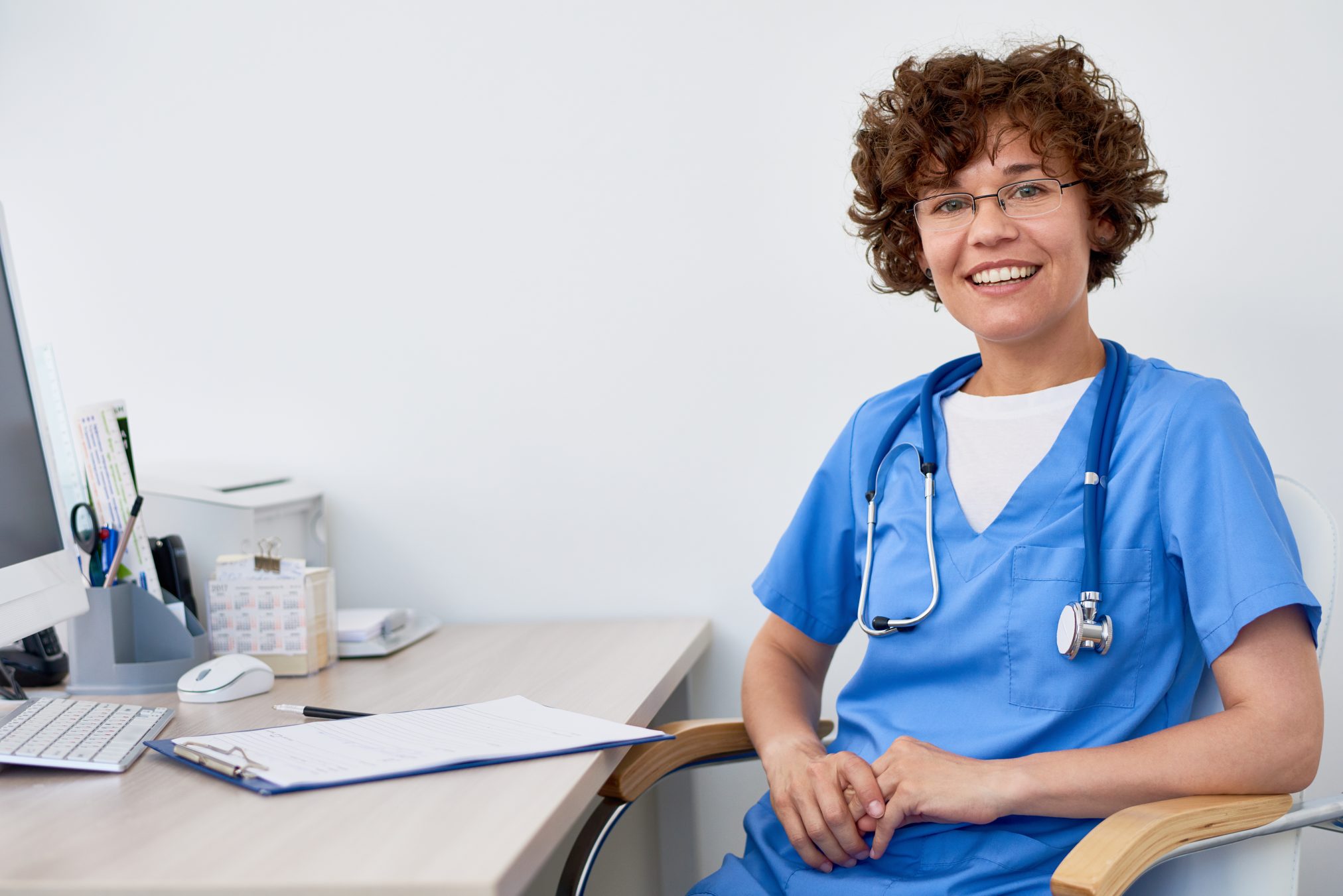 Portrait of friendly female doctor sitting at desk in office , posing smiling happily at camera